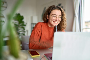 Looking for an educational experience that’s more than lectures and tests? You may be interested in CTE –– Career and Technical Education. This is a gateway to real world skills and a head start on the career of your dreams. -Happy young woman using laptop sitting at desk writing notes while watching webinar, studying online, looking at pc screen learning web classes or having virtual call meeting remote working from home.